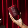 Hand holding a red candle with a dark label against a red curtain background